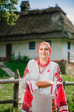 a Ukrainian woman in traditional attire holds rushnyk with bread and salt on the background of an old house and flowers