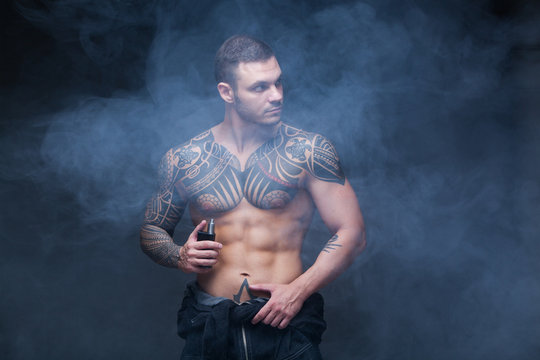 Vaper. The man with a muscular naked torso with tattoos smoke an electronic cigarette on the dark background