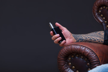 Hand close up. Vaper. The man with tattoos sits on a leather sofa smoke an electronic cigarette on...