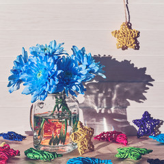 A bouquet of blue chrysanthemums is in a glass jar on a white wooden background. Wicker colorful figures of stars and crescents are scattered nearby.