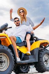 Naklejka premium happy young couple sitting on ATV in front of cloudy sky and looking at camera