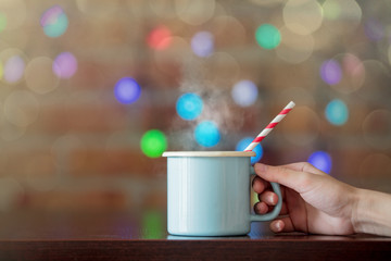 Female hand holding a hot cup of coffee with fairy lights on background. Christmas season