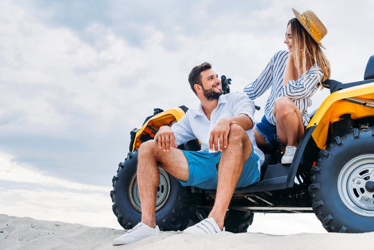 happy young couple sitting on ATV on sandy dune in front of cloudy sky