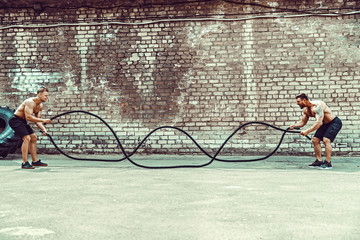Two muscular athletes training, working out with rope in front of brick wall. Scream. Working hard. Street gym. Strength and motivation. Outdoor workout. © zamuruev