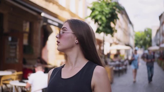 Pretty young Caucasian woman wearing stylish sunglasses walking through the street, touching hair. Good-looking Slavic girl looking around. Summer in big city. Outdoors.