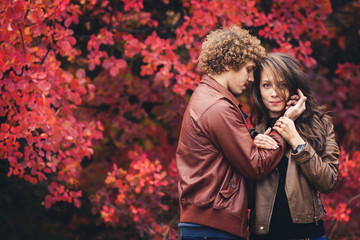 Curly-haired mustachioed man and brown-haired woman hugging in autumn against background of red trees.