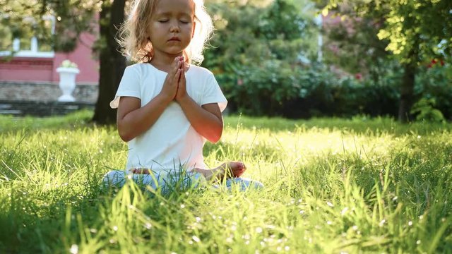 Little cute baby girl meditates on the grass at green summer park.