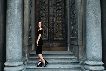 Fototapeta na wymiar Cute young girl modeling appearance posing against the backdrop of huge beautiful doors in black dress and hat