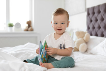 Adorable little baby with book on bed at home