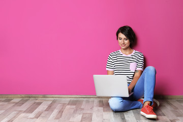 Young woman with modern laptop sitting on floor near color wall