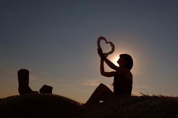 A young woman dressed as a cowgirl holds a straw heart in her hand. She takes time out and enjoys the summer sun  setting as she leisures on a hay bale.