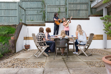 Father and daughter playing their guitars to family members on the patio area of their garden.
