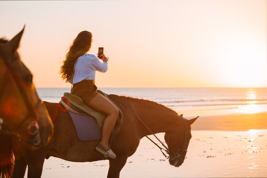 Young woman taking sunset photos while riding a horse at the bea
