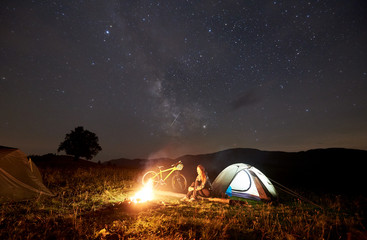Young woman traveller enjoying at night camping near burning campfire, illuminated tourist tent, mountain bike under beautiful evening sky full of stars. Outdoor activity and tourism concept
