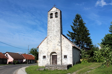 Fototapeta na wymiar Stone church with tall stone bell tower next to paved road and small family houses surrounded with uncut grass with cloudy blue sky in background