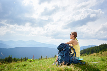Fototapeta na wymiar Back view of attractive woman tourist resting on grassy hill with backpack and trekking sticks. Female backpacker enjoying summer cloudy day in the mountains. Outdoor activity, tourism concept