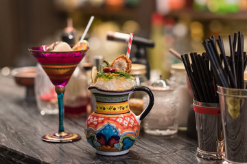Fancy cocktails in red patterned wineglass and original painted jar are standing on bar counter with cocktail tools and ingredients. Modern trendy alcoholic drinks in club. Party background concept.
