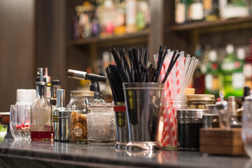 Professional cocktail tools and ingredients in glass bottles and jars for creative modern fashionable cocktails on wooden bar counter. Blurred bottles with alcoholic drinks on background. 