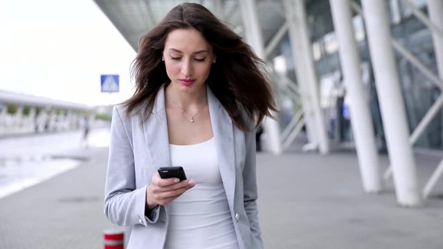 Charming Young Woman Walking with her Smartphone. Attractive Girl Typing a Message While Walking in the City. Looking Stylish. Wearing Classical Suit. Neat Hairstyle. Businesswoman Resting.