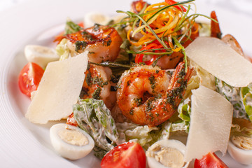 Caesar salad with quail eggs, cherry tomatoes, Parmesan cheese and grilled shrimps in white plate on light table.