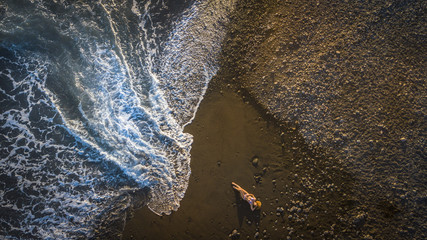 beautiful cacuasian woman model laying down on the black vulcanic beach and taking sun while a big wave is coming. outdoor nature and lifestyle concept. vacation and travel image. aerial view.