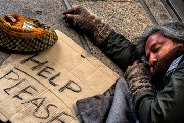 homeless male getting to sleep on floor in public path way with old hat in front for money donation
