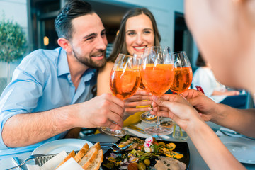 Handsome Middle-Eastern young man sitting next to his Caucasian girlfriend while toasting with two mutual friends outdoors at a trendy restaurant in summer