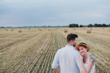 Happy stylish cheerful caucasian couple walking and spending time together in summer wheat field. Man and woman hugging and kissing outdoors. Love, romance, happiness concept.