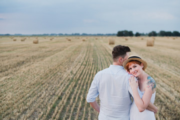 Happy stylish cheerful caucasian couple walking and spending time together in summer wheat field. Man and woman hugging and kissing outdoors. Love, romance, happiness concept.