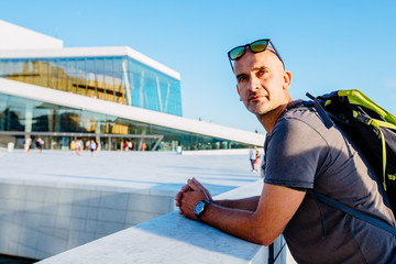 Pensive handsome middle age man traveler with backpack standing and looking away over Oslo Opera House urban background. - obrazy, fototapety, plakaty