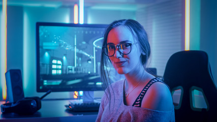 Portrait of the Beautiful Young Pro Gamer Girl Sitting at Her Personal Computer and Looks into Camera. Attractive Geek Girl Player Wearing Glasses in the Room Lit by Neon Lights. - obrazy, fototapety, plakaty