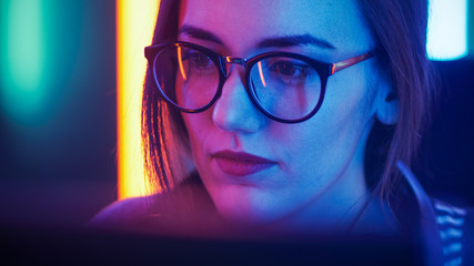 Portrait of the Beautiful Young Girl Sitting Before Computer, Browsing in Internet, Playing Online Games, Streaming. Cute Girls Wearing Glasses in the Cool Retro Neon Lit Room.