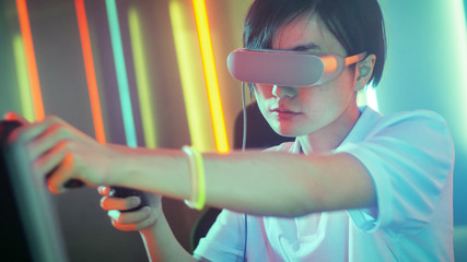 East Asian Pro Gamer Wearing Virtual Reality Headset Plays Online Video Game with Joysticks / Controllers. Cool Retro Red Neon Colors in the Room. © Gorodenkoff