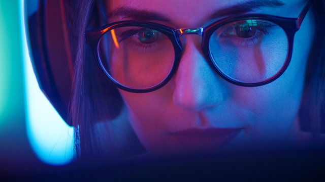 Portrait of the Beautiful Young Girl Sitting Before Computer, Browsing in Internet, Playing Online Games, Streaming. Cute Girls Wearing Glasses in the Cool Retro Neon Lit Room.