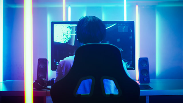 Back View Shot of the Professional Gamer Playing in First-Person Shooter Online Video Game on His Personal Computer. Room Lit by Neon Lights in Retro Arcade Style. Online Cyber e-Sport Championship.