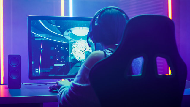 Back View Shot of the Beautiful Professional Gamer Girl Playing in Online Shooter Online Video Game on Her Personal Computer. Casual Cute Geek Girl Wearing Headset. Lit by Neon Lights in Retro Style.