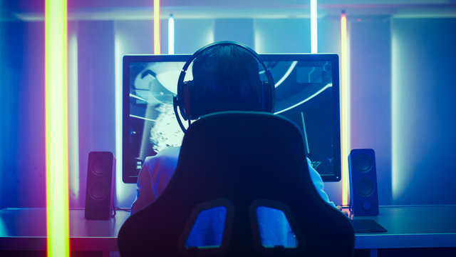 Back View Shot of the Professional Gamer Playing in First-Person Shooter Online Video Game on His Personal Computer. Room Lit by Neon Lights in Retro Arcade Style. Online Cyber e-Sport Internet.