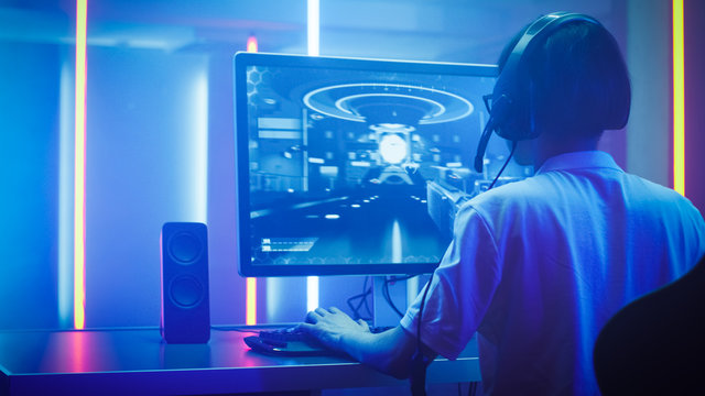 Arc Shot of the Professional Gamer Playing in First-Person Shooter Online Video Game on His Personal Computer. He's Talking with His Team Through Headset. Room Lit by Neon Lights in Retro Arcade Style
