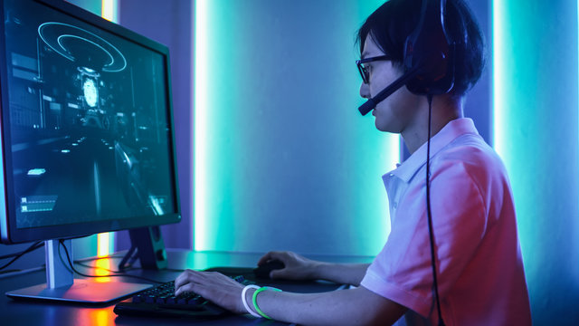Professional East Asian Gamer Playing in First-Person Shooter Online Video Game on His Personal Computer. Gameplay Walkthrough Showing on the Screen. Room Lit by Neon Lights in Retro Arcade Style