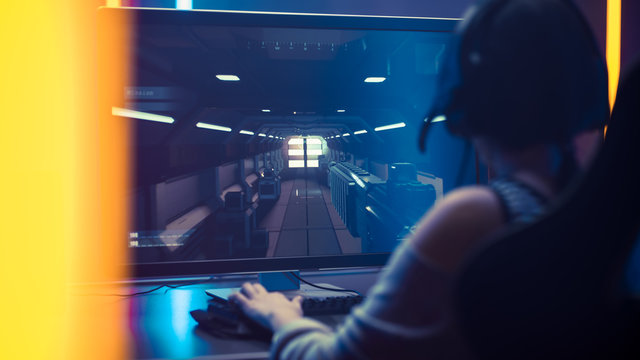 Shot of the Beautiful Pro Gamer Girl Playing in First-Person Shooter Online Video Game on Her Personal Computer. Casual Cute Geek wearing Glasses and Using Headset. Neon Room.