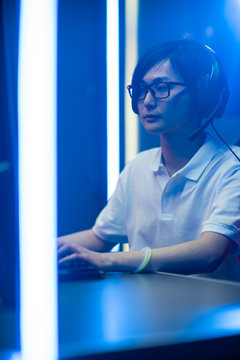 Professional Gamer Playing Online Video Game on His Personal Computer. He's Talking with His Team Through Headset. Room Lit by Neon Lights in Retro Arcade Style. Vertical Shot.