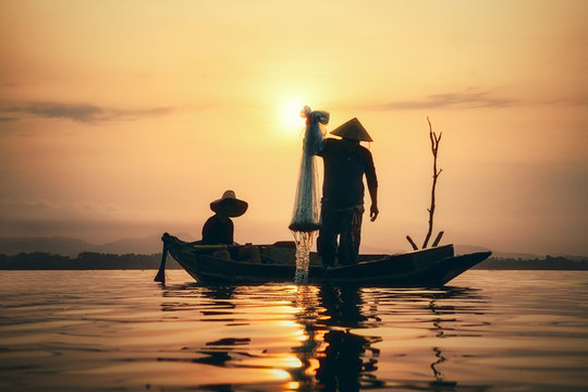 Fisherman on wooden boat casting a net for catching freshwater fish in reservoir at sunrise.