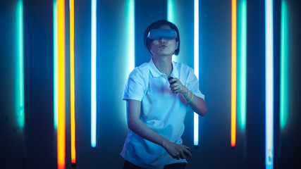 East Asian Pro Gamer Wearing Virtual Reality Headset Plays Online Video Game Shooter using Joysticks / Controllers as Swords. Cool Retro Neon Colors in the Room.