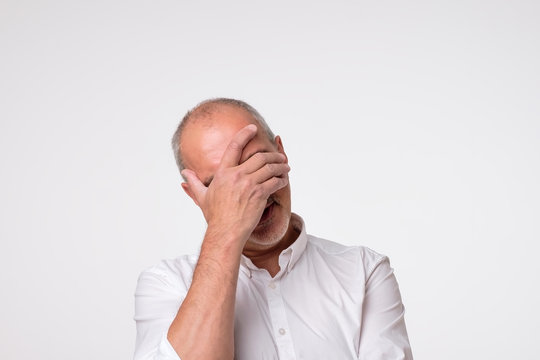 Displeased mature man in white shirt covering his eyes over gray background