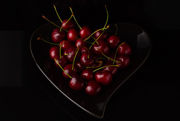 Cherries on black plate in form of heart. Cherry berry on a black background. Dark image of red cherries on a black reflective surface.