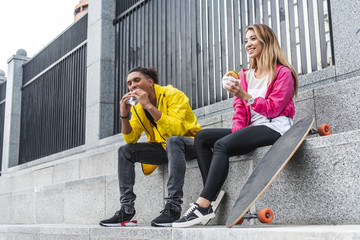young multicultural couple of skateboarders eating burgers at city street