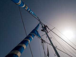 Skipper climbs up on the pole of a sail boat. View from deck up to sky.