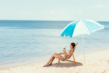 Fototapeta na wymiar beautiful girl in bikini with coconut cocktail resting on deck chair under beach umbrella near sea