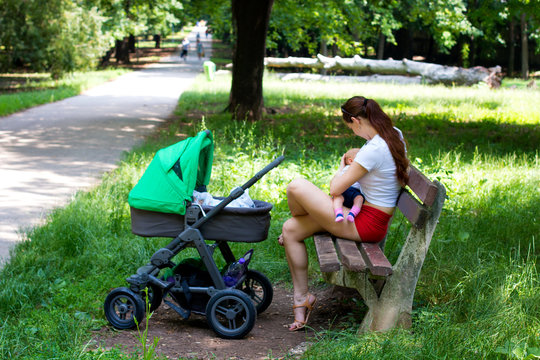 Woman parent is sitting on the wooden bench in the park, holding and breastfeeding baby, next to stroller, public park with green grass during sunny day, mother and infant