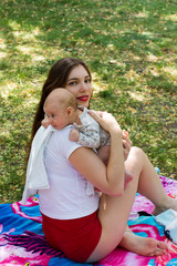 Happy mother is caring a baby, infant head is resting on mom's shoulder, pretty woman and his beautiful baby in embrace, sitting on the blanket at the park, warm summer day